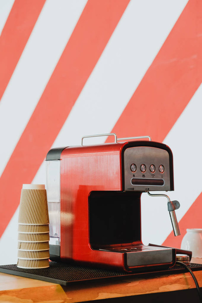 A nice shiny red coffee machine with paper and ceramic cups on  a stripy background wall. - Photo, Image