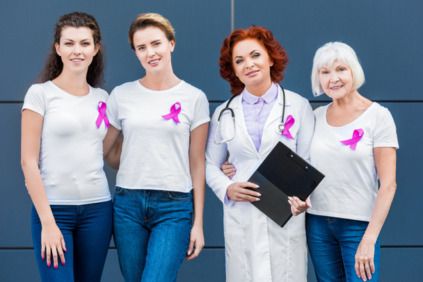 women and doctor with pink ribbons standing together and smiling at camera, breast cancer awareness concept - Photo, Image