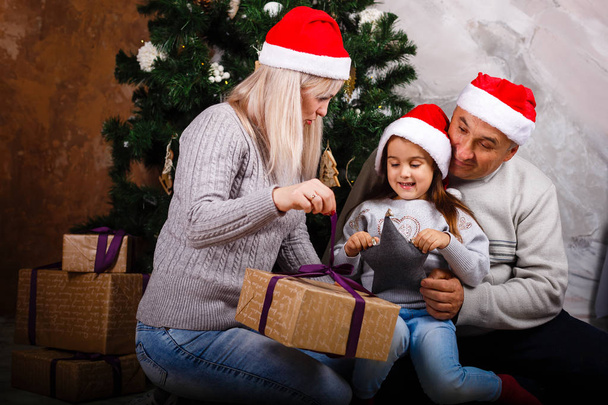 kleines Mädchen öffnet Geschenkschachtel, während Mama und Großvater zu Hause am Weihnachtsbaum sitzen  - Foto, Bild
