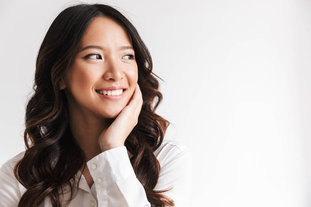 Image of smiling chinese woman with long dark hair looking aside at copyspace and touching cheek isolated over white background in studio - Foto, imagen