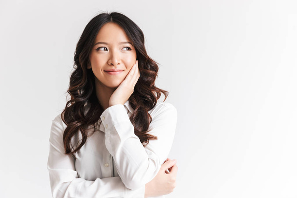 Portrait of happy chinese woman with long dark hair looking aside at copyspace and touching cheek isolated over white background in studio - 写真・画像