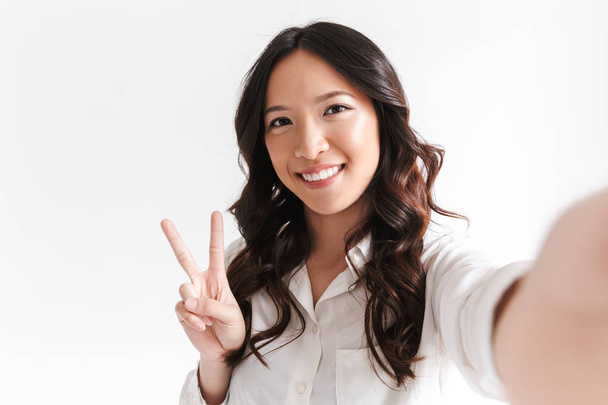 Portrait of optimistic chinese woman with long dark hair smiling and taking selfie photo with peace sign isolated over white background in studio - Photo, image