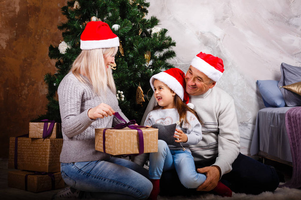 kleines Mädchen öffnet Geschenkschachtel, während Mama und Großvater zu Hause am Weihnachtsbaum sitzen  - Foto, Bild