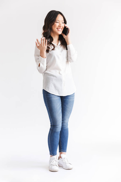 Full length photo of joyous chinese woman with long dark hair holding and talking on mobile phone isolated over white background in studio - Foto, afbeelding