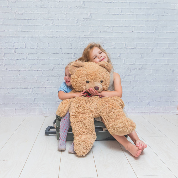 two girls are sitting on a suitcase with a toy bear on the background of a white brick wall - Foto, imagen