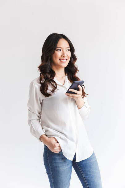 Foto de mujer asiática feliz con pelo largo y oscuro sosteniendo el teléfono móvil negro y mirando a un lado aislado sobre fondo blanco en el estudio
 - Foto, Imagen