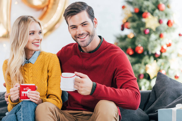 smiling couple with cups of coffee at home on christmas eve - Fotografie, Obrázek