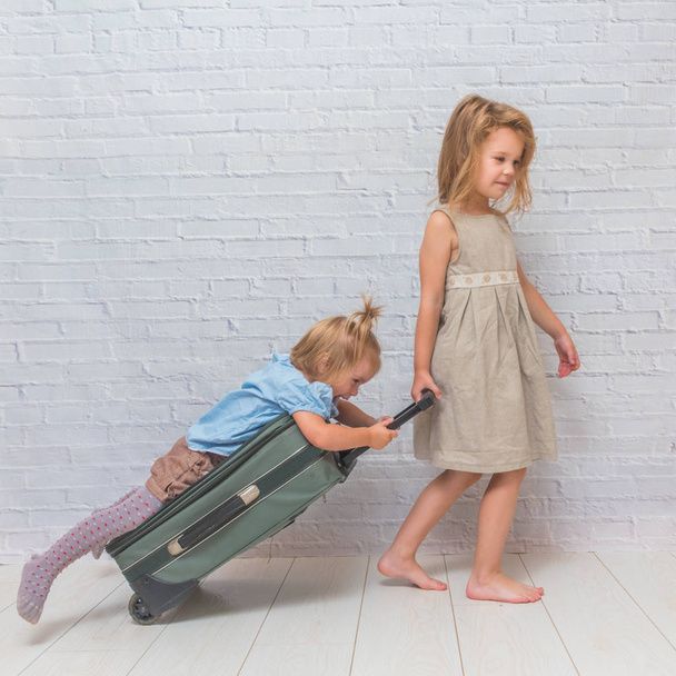 a two girls, kids with suitcase on wheels on white brick wall background - Zdjęcie, obraz