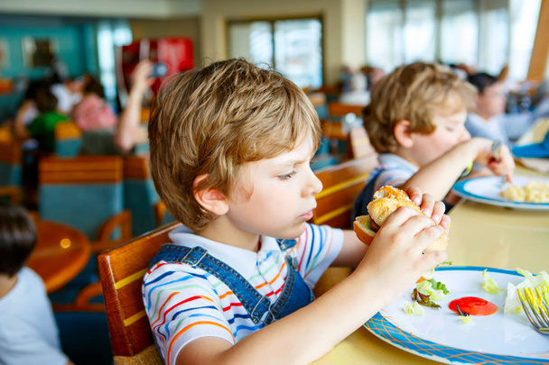Cute healthy preschool kid boy eats hamburger sitting in school or nursery cafe. Happy child eating healthy organic and vegan food in restaurant. Childhood, health concept. - Photo, Image