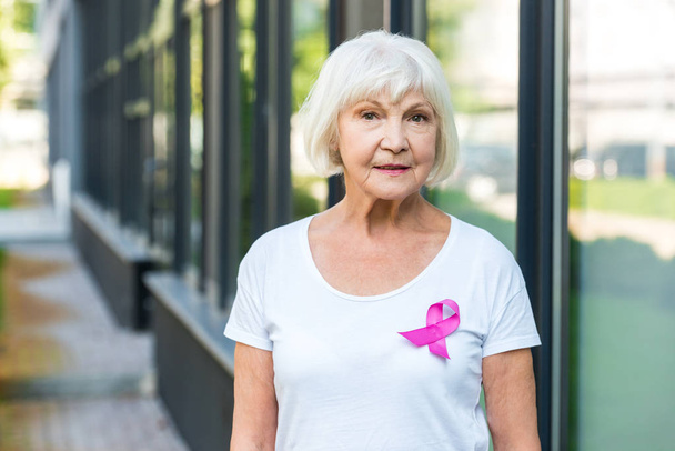 senior woman with pink ribbon on t-shirt looking at camera, breast cancer awareness concept       - Фото, зображення