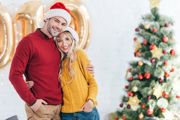 счастливая молодая пара в шляпах Санты обнимается дома с рождественской ёлкой
 - Фото, изображение