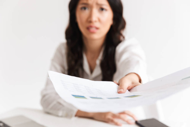 Blurry photo of tense asian woman employee with long dark hair holding and giving papers at camera while working in office isolated over white background - Foto, immagini