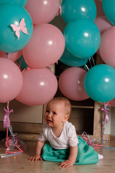 a little girl, a child, sits on the floor and cries, wrinkled her nose, near the balloons, a holiday. birthday, turned a year old. dress is white and blue. resentment, disappointment.the child cries, gets angry.frustrated baby. cry - Foto, Imagen