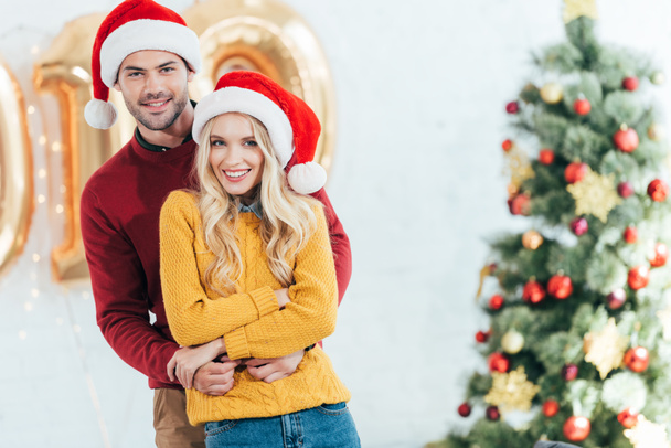 happy couple in santa hats embracing at home with christmas tree - Fotó, kép