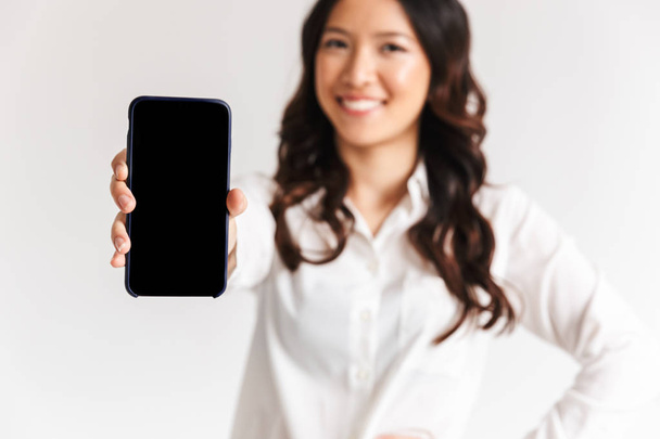 Blurry photo of smiling asian woman with long dark hair standing and showing smartphone at camera isolated over white background in studio - Foto, Imagem