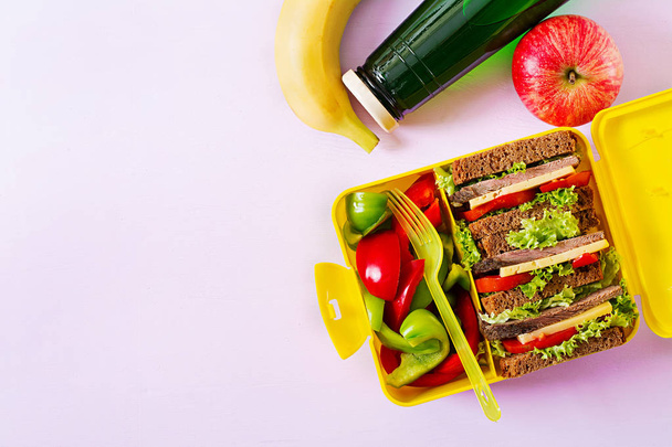 Healthy school lunch box with beef sandwich and fresh vegetables, bottle of water and fruits on pink background. Top view. Flat lay - Photo, Image