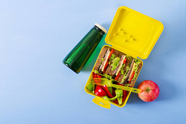 Healthy school lunch box with beef sandwich and fresh vegetables, bottle of water and fruits on blue background. Top view. Flat lay - Photo, Image
