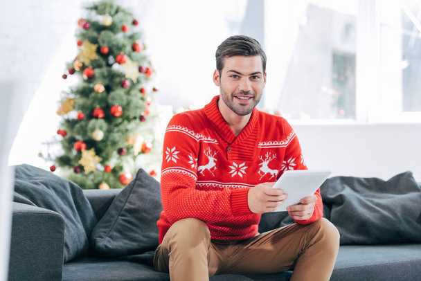 happy man using digital tablet and sitting on sofa with christmas tree behind - Фото, изображение