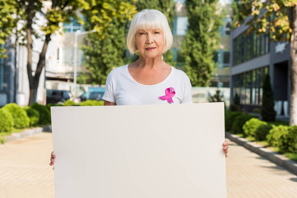 空白のカードを持っているとカメラ、乳房癌意識概念を見てピンクのリボンと年配の女性  - 写真・画像