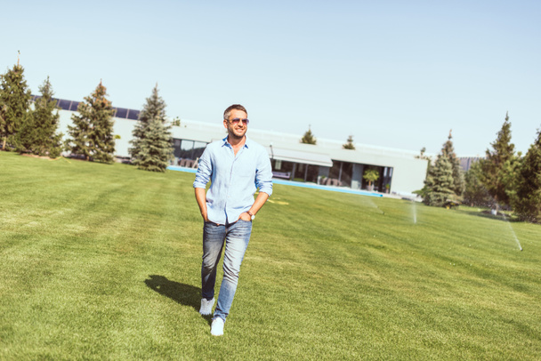 handsome adult man in sunglasses walking with hands in pockets on lawn near country house  - Fotoğraf, Görsel
