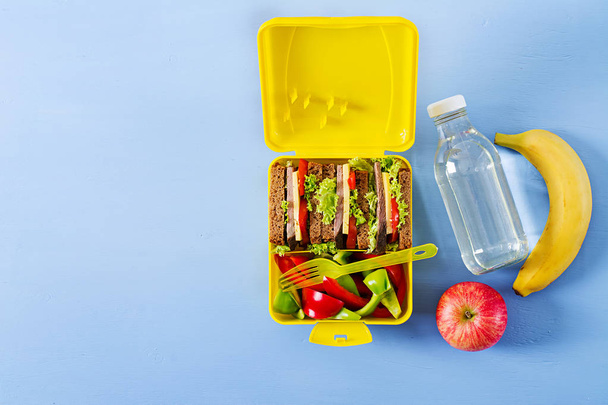 Healthy school lunch box with beef sandwich and fresh vegetables, bottle of water and fruits on blue background. Top view. Flat lay - Foto, imagen