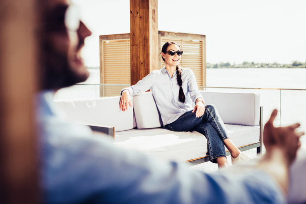 selektiver Fokus einer lächelnden Frau mit Sonnenbrille auf dem Sofa, während ihr Freund in der Nähe im Landhaus sitzt  - Foto, Bild