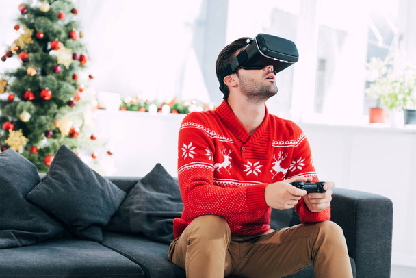 Человек с гарнитурой виртуальной реальности и джойстиком играет в видеоигру накануне Рождества
 - Фото, изображение