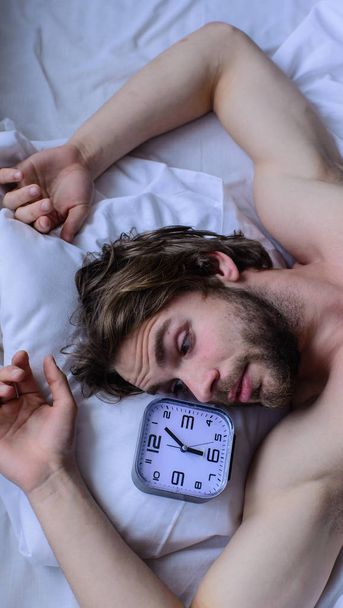 Toughest part of morning simply getting out of bed. Get up early morning tips. Man bearded unshaven handsome guy lay on pillow near alarm clock top view. Guy relaxing bed before alarm clock ringing - 写真・画像