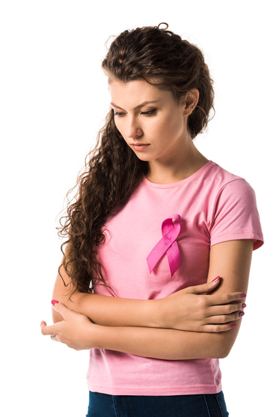 νεαρή γυναίκα με κορδέλα ροζ στέκεται με σταυρωμένα τα χέρια και κοιτάζοντας προς τα κάτω απομονωμένα σε λευκό, έννοια του καρκίνου μαστού  - Φωτογραφία, εικόνα