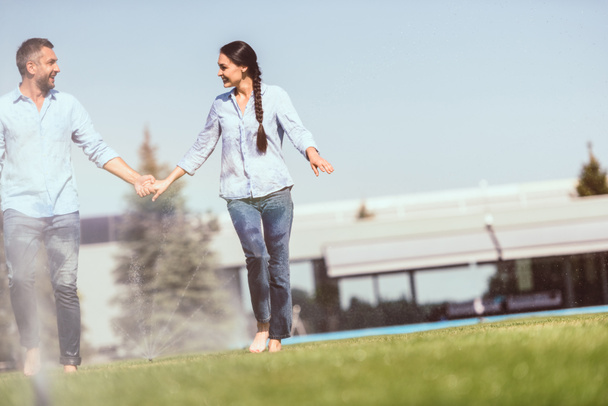 happy couple holding hands and having fun with automatic watering on grass near country house  - Fotografie, Obrázek