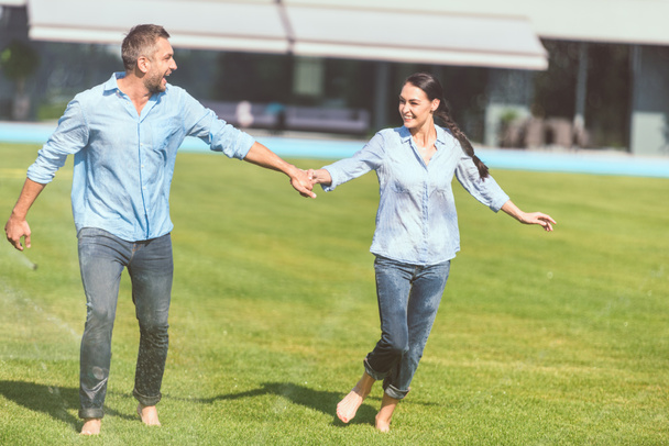 усміхнена пара тримає руки і розважається з автоматичним поливом на зеленому газоні біля заміського будинку
  - Фото, зображення