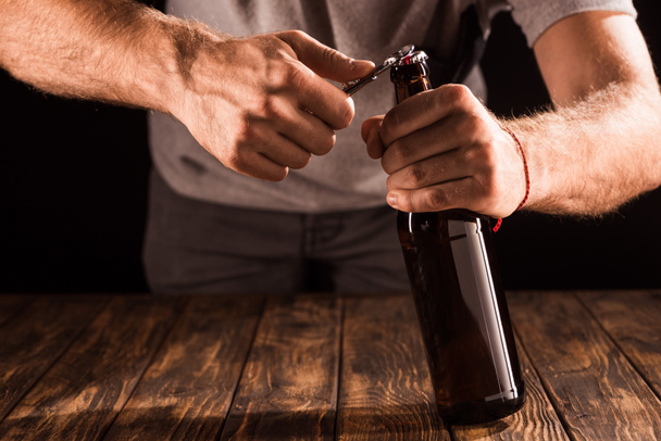木製のテーブルで開幕戦でビール瓶を開く男の部分的なビュー  - 写真・画像