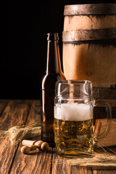 mug of fresh beer with foam, beer bottle, wheat and wooden barrel at table on black background - Foto, imagen