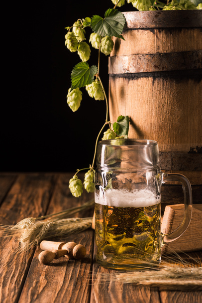 Mok verse bier met schuim, tarwe, hop en houten vat aan tafel op zwarte achtergrond - Foto, afbeelding