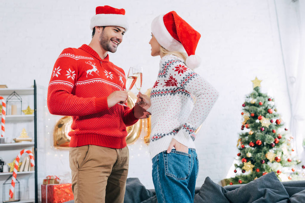 junges Paar in Weihnachtsmützen prostet mit Sektgläsern zu Hause mit Weihnachtsbaum zu - Foto, Bild