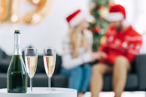 enfoque selectivo de botella de champán y vasos para celebrar la Navidad, pareja sentada detrás
 - Foto, Imagen