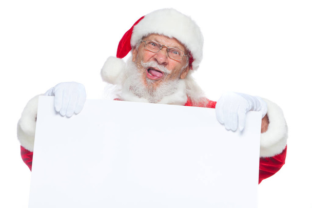 Christmas. Evil Santa Claus in white gloves with his tongue sticking out holds an empty white cardboard and shows faces, grimaces. Place for advertising, for text, empty space. Copy-paste. Isolated on - Photo, Image