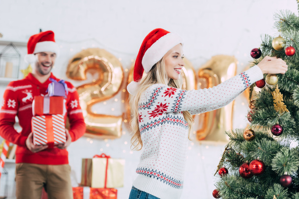 attractive woman decorating christmas tree while happy man carrying gift boxes - Photo, Image