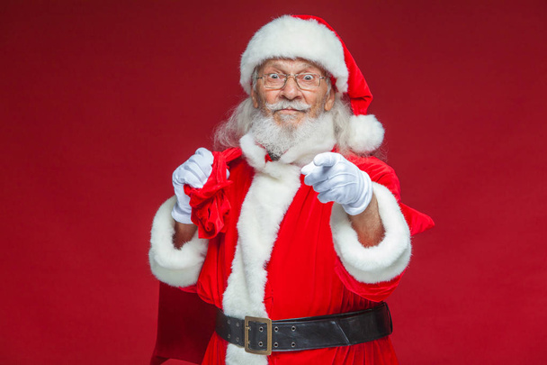 Christmas. Smiling Santa Claus in white gloves with a bag of gifts behind him points his index finger into the camera. Isolated on red background. - Fotoğraf, Görsel