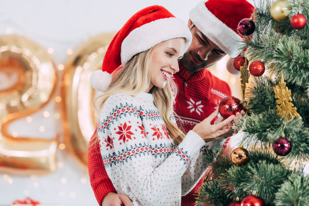 young couple in santa hats decorating christmas tree together - Valokuva, kuva