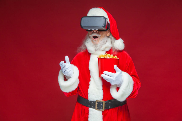 Christmas. Santa Claus in black virtual reality glasses holding a red bucket of popcorn. Watching a movie in virtual reality. Isolated on red background. - Fotó, kép