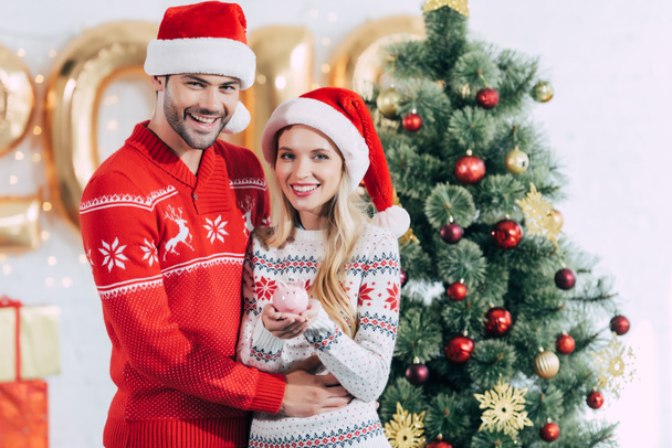счастливая пара в шляпах Санты, держащая копилку с банкнотой дома с рождественской елкой
 - Фото, изображение