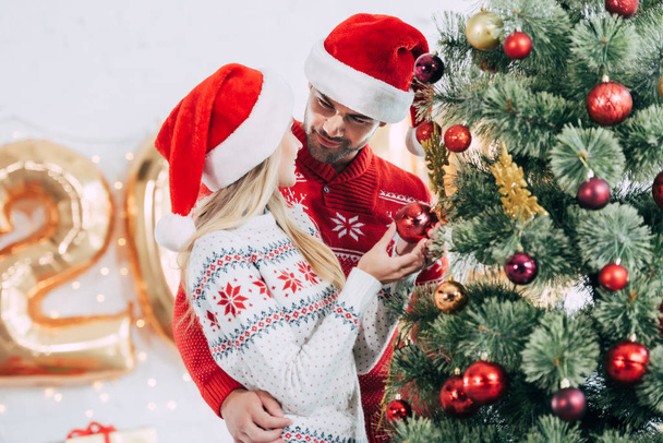happy couple in santa hats decorating christmas tree at home together - Foto, Imagem