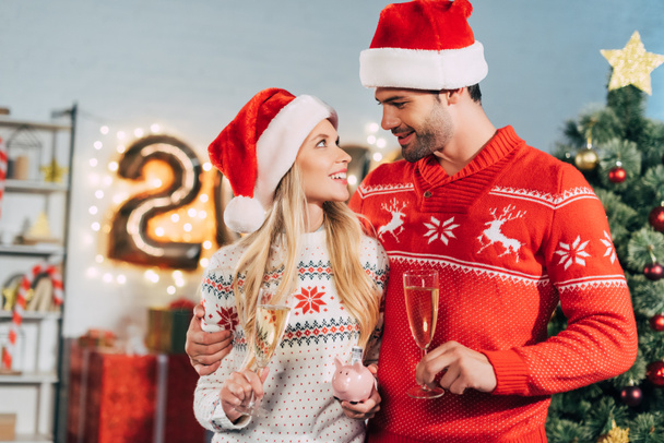 пара в шляпах Санта-Клауса с бокалами для шампанского, держащая копилку со сбережениями на Рождество
 - Фото, изображение