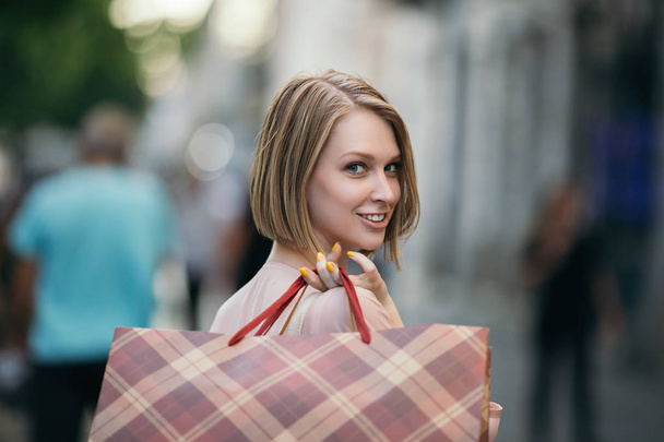Beautiful and fashionable young woman with shopping bags standing on city street and looking at shopfront - Foto, Bild