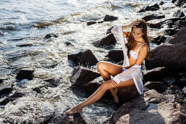Portrait of beautiful brunette woman in sunlight. Model in white lingerie on rocky beach at daytime - Фото, зображення