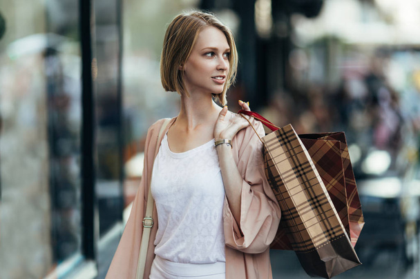 Beautiful and fashionable young woman with shopping bags standing on city street and looking at shopfront - Fotoğraf, Görsel