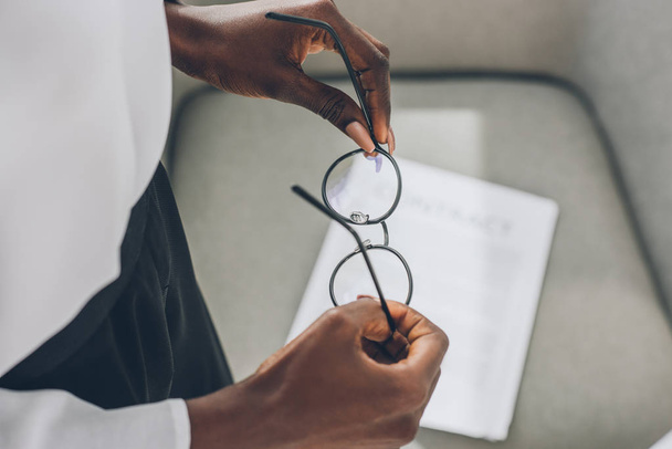 imagem recortada de empresária afro-americana segurando óculos perto de poltrona no escritório
 - Foto, Imagem
