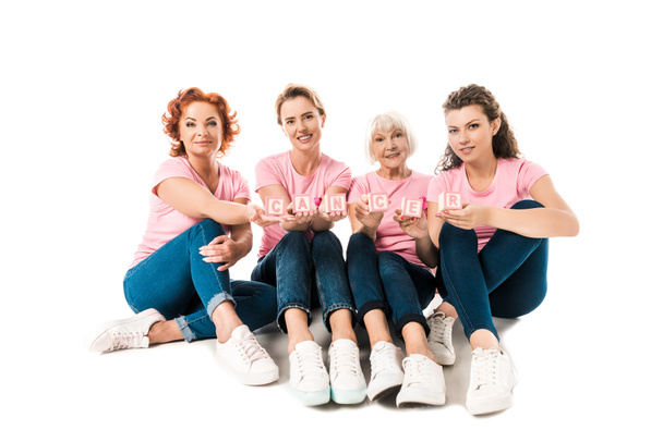 women in pink t-shirts holding cubes with word cancer and smiling at camera while sitting together isolated on white - Photo, Image