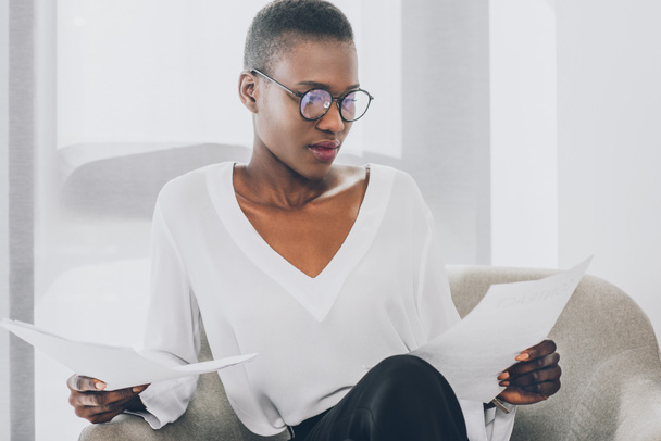 stylish attractive african american businesswoman sitting in armchair and reading documents in office - Фото, изображение